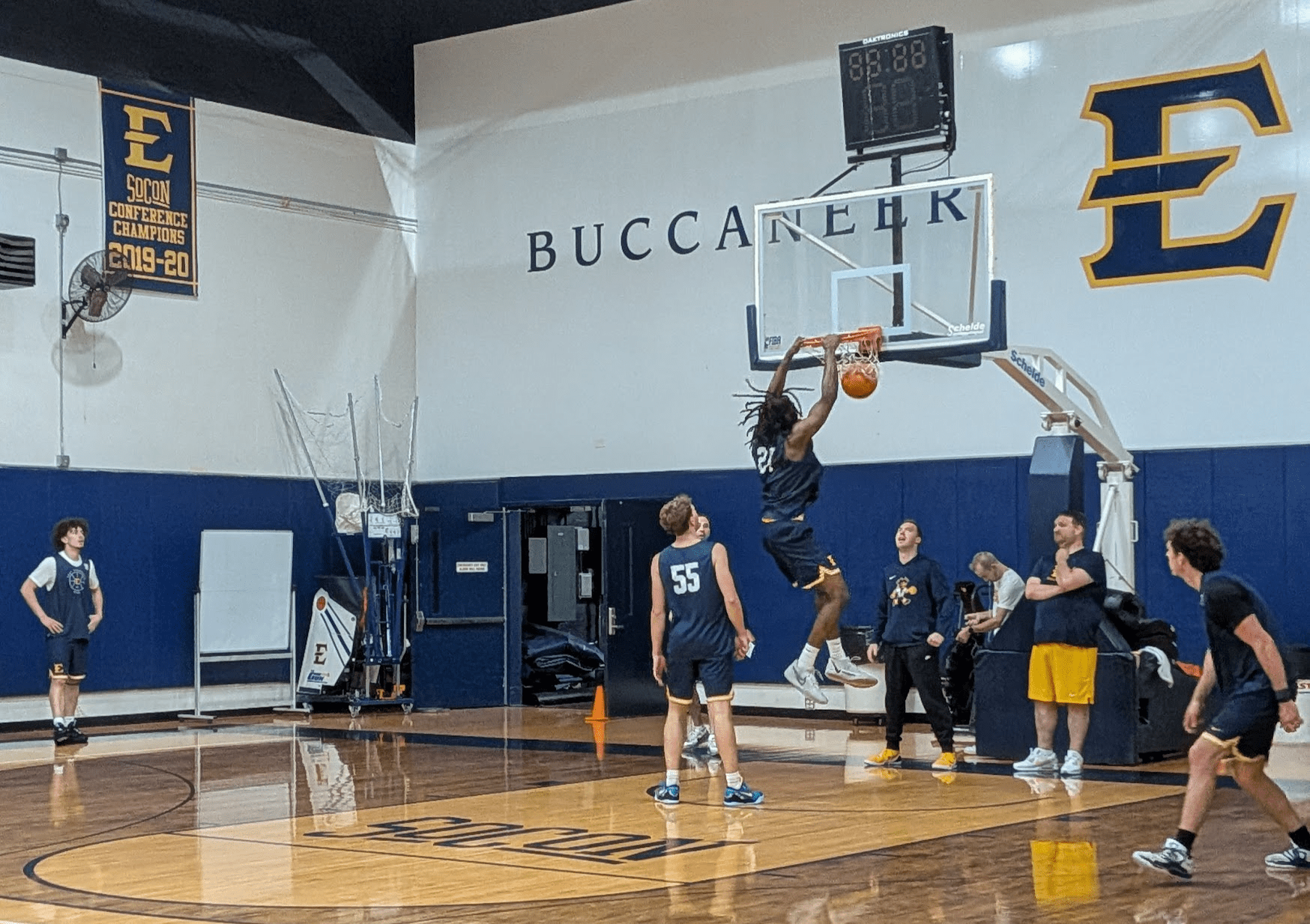 Player in a blue uniform dunks the ball during practice in a gym with 'BUCCANEER' lettering on the wall.