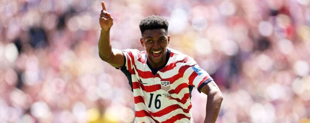 Soccer player in a red-and-white striped USA jersey with number 16 celebrates, pointing upward on the field.