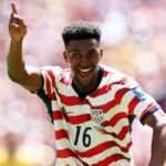 Soccer player in a red-and-white striped USA jersey with number 16 celebrates, pointing upward on the field.