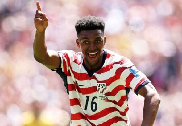 r1676775_2_1296x518_5-2 » Greenville Sports Media Soccer player in a red-and-white striped USA jersey with number 16 celebrates, pointing upward on the field.