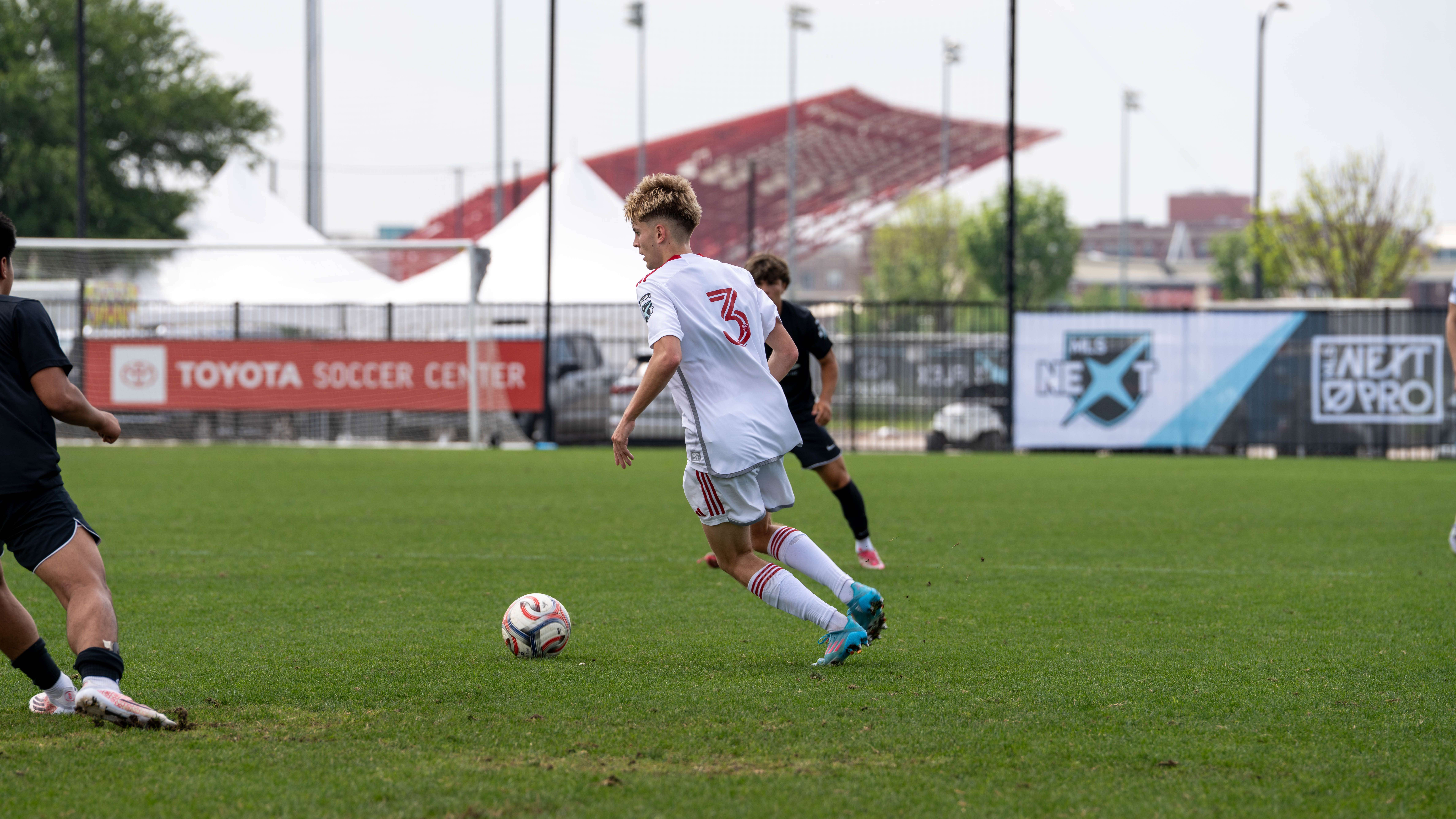 Soccer player in a white jersey dribbles a ball on a green field while teammates and opponents watch, with banners and stadium seating in the background.