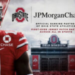 Ohio State football player in red jersey with sponsor logos, standing before Ohio Stadium banner.