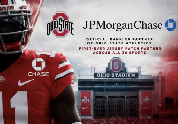 Ohio State football player in red jersey with sponsor logos, standing before Ohio Stadium banner.