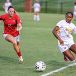 Two female soccer players chase a ball on a green field during a match; player in red (number 12) is ahead.