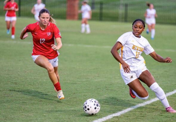 Two female soccer players chase a ball on a green field during a match; player in red (number 12) is ahead.