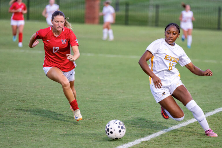 Two female soccer players chase a ball on a green field during a match; player in red (number 12) is ahead.