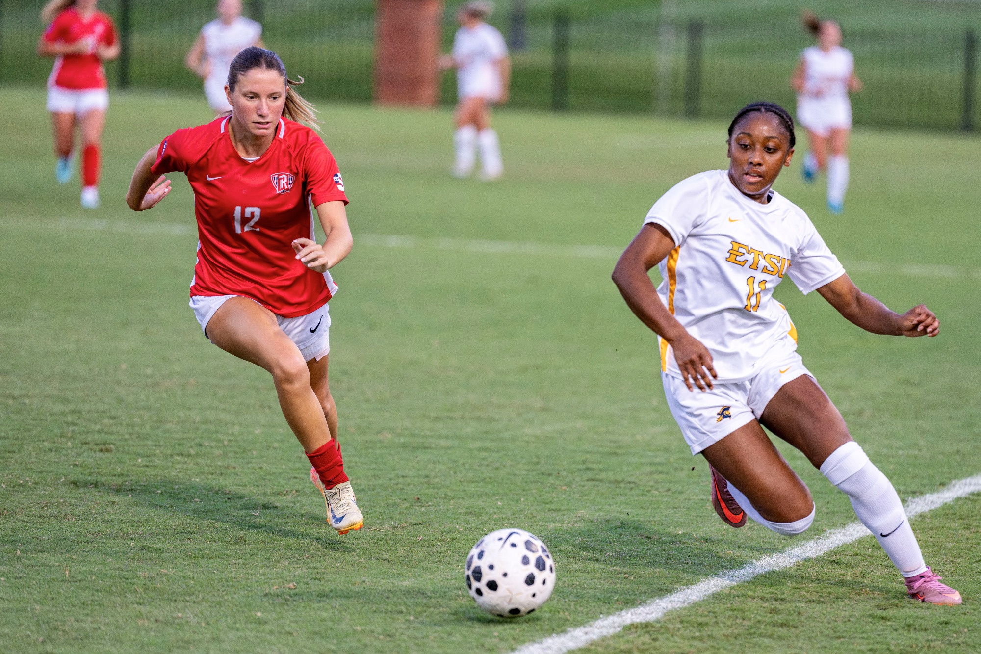 Two female soccer players chase a ball on a green field during a match; player in red (number 12) is ahead.