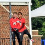 Two goalkeepers in red jerseys stand by a soccer goal, brick wall behind the net, outdoors on a sunny day.