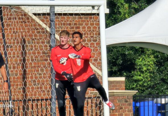 Two goalkeepers in red jerseys stand by a soccer goal, brick wall behind the net, outdoors on a sunny day.