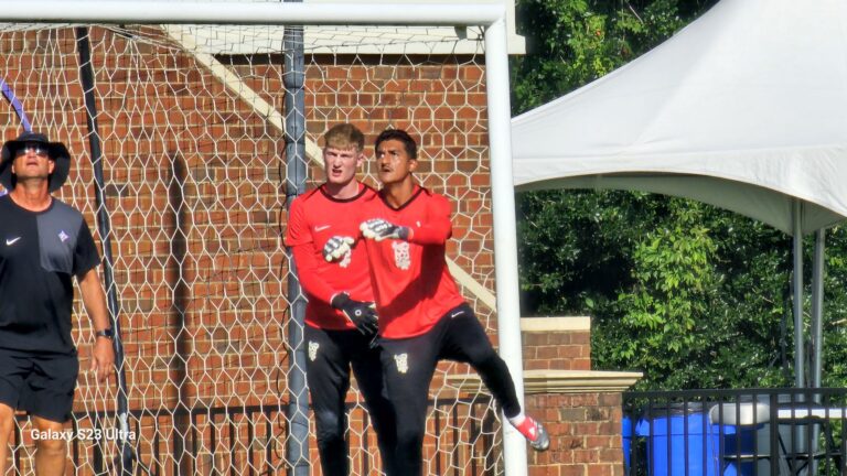 Two goalkeepers in red jerseys stand by a soccer goal, brick wall behind the net, outdoors on a sunny day.