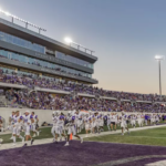College football team in white uniforms runs onto a purple end zone as sunset lights up the stadium stands filled with fans.