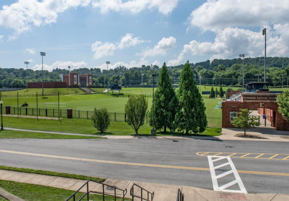 Wide view of a university athletic field and brick stadium buildings under a blue sky with tall light towers and green grass surrounding the field.
