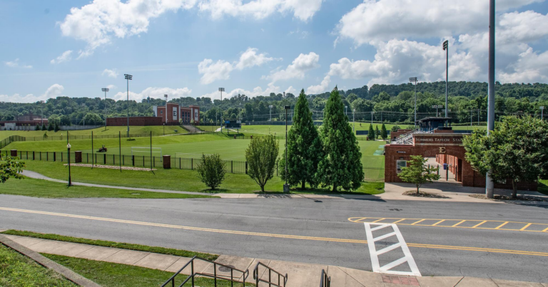 Wide view of a university athletic field and brick stadium buildings under a blue sky with tall light towers and green grass surrounding the field.