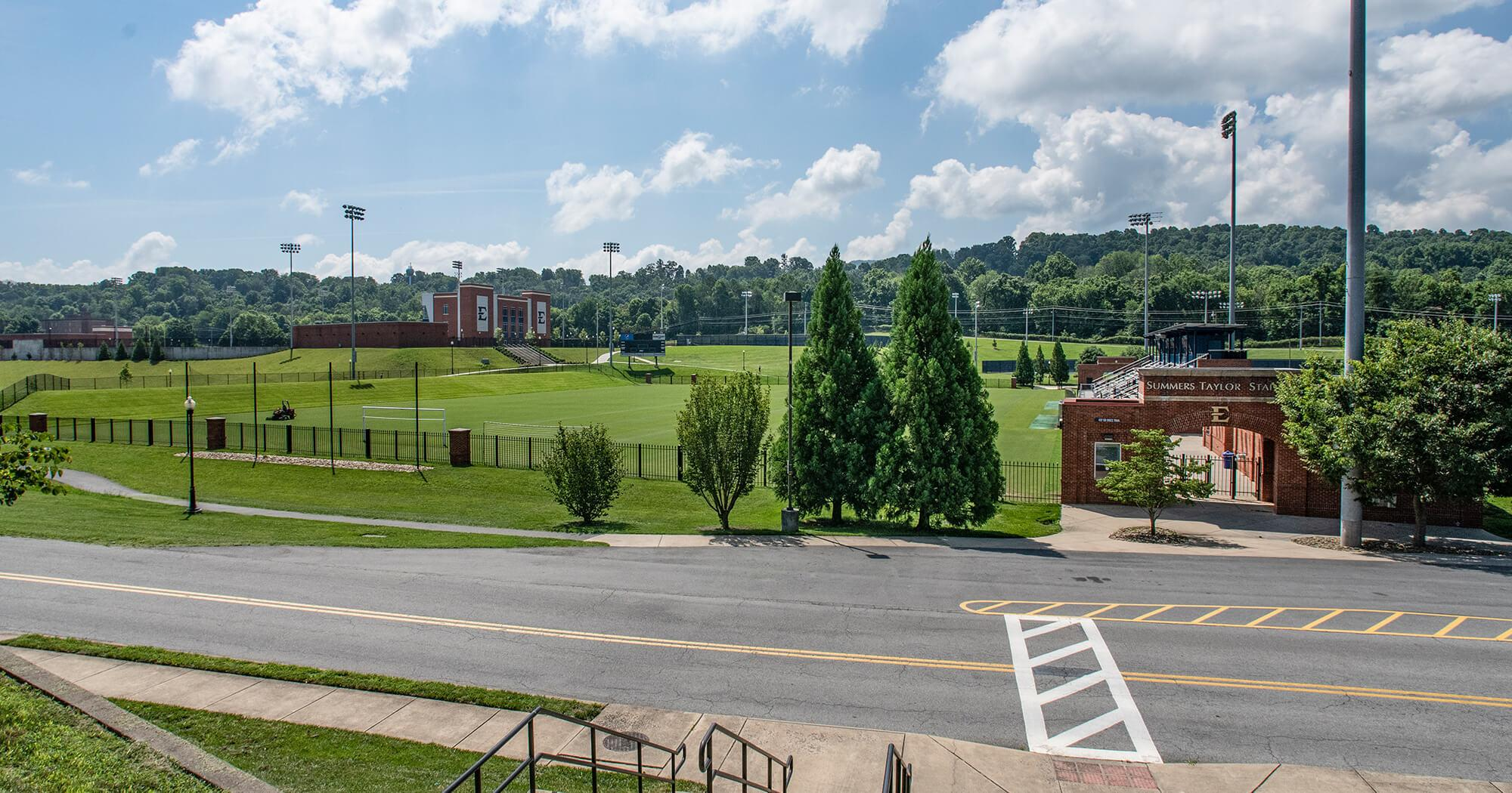 Wide view of a university athletic field and brick stadium buildings under a blue sky with tall light towers and green grass surrounding the field.