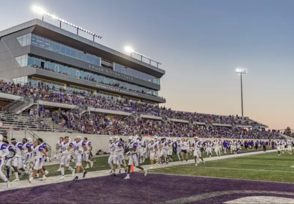 College football team in white uniforms runs onto a purple end zone as sunset lights up the stadium stands filled with fans.