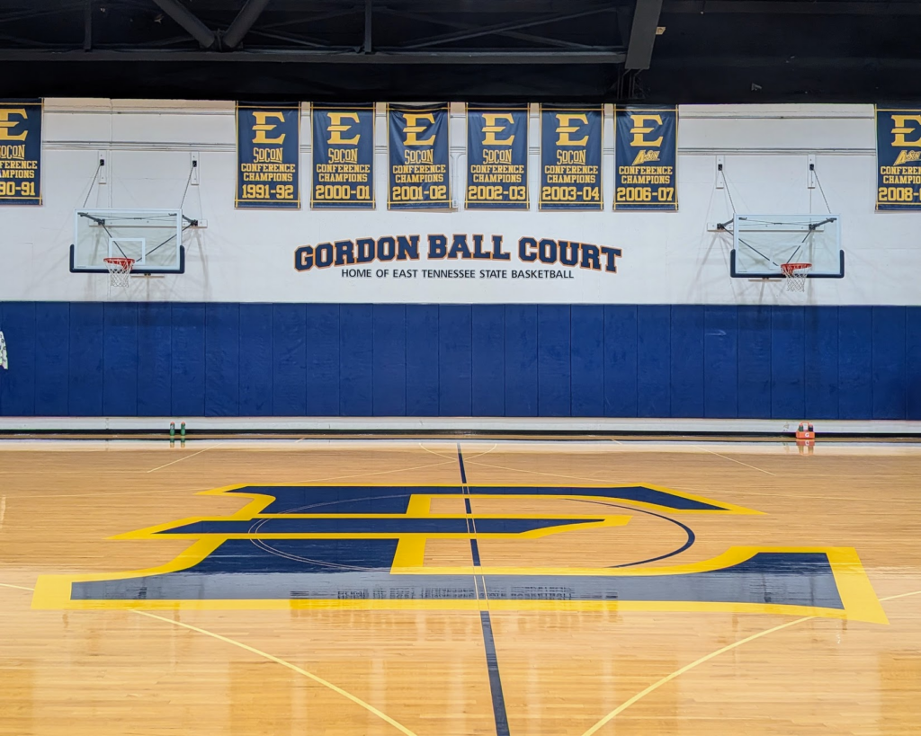 Empty basketball gym with two hoops, blue padded wall, and championship banners overhead, centered by a blue-yellow court logo saying 'Gordon Ball Court'