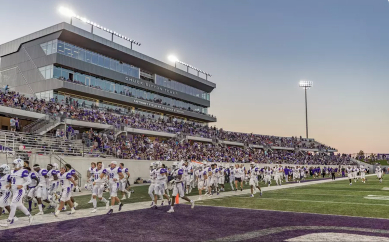 College football team in white uniforms runs onto a purple end zone as sunset lights up the stadium stands filled with fans.